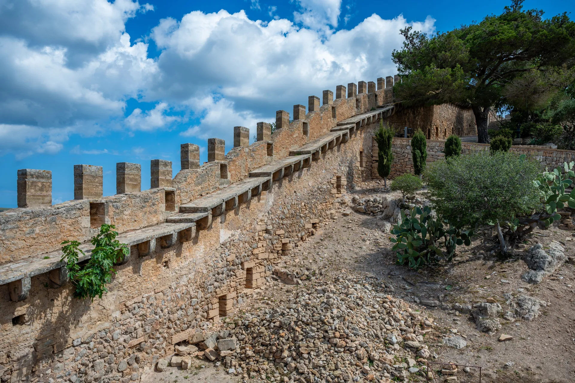 Muraille médiévale en pierre avec des créneaux sous un ciel bleu parsemé de nuages blancs, entourée de végétation méditerranéenne