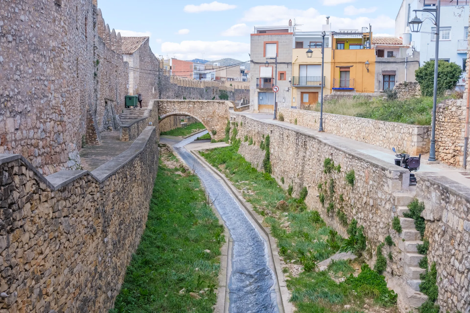 Un canal d'eau étroit bordé de murs en pierre et de verdure, avec des bâtiments colorés et un pont en arrière-plan sous un ciel nuageux.