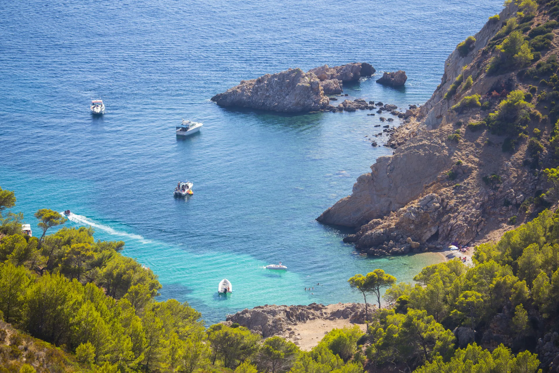 Vue aérienne d'une crique rocheuse avec des eaux turquoise et des bateaux, entourée de pins verts luxuriants sous un ciel ensoleillé.