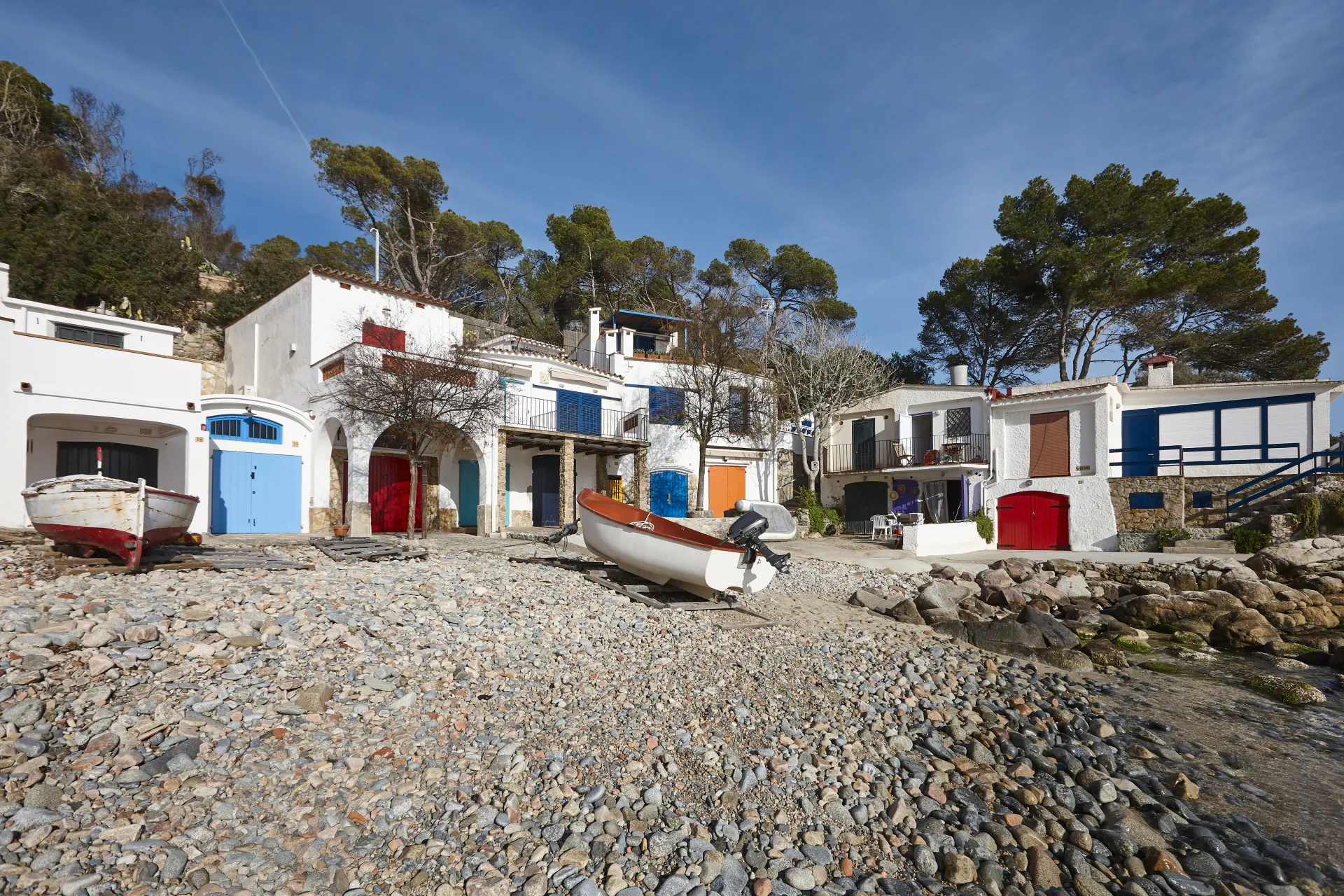 Maisons blanches traditionnelles avec des portes colorées et des bateaux de pêche sur une plage de galets sous un ciel bleu clair