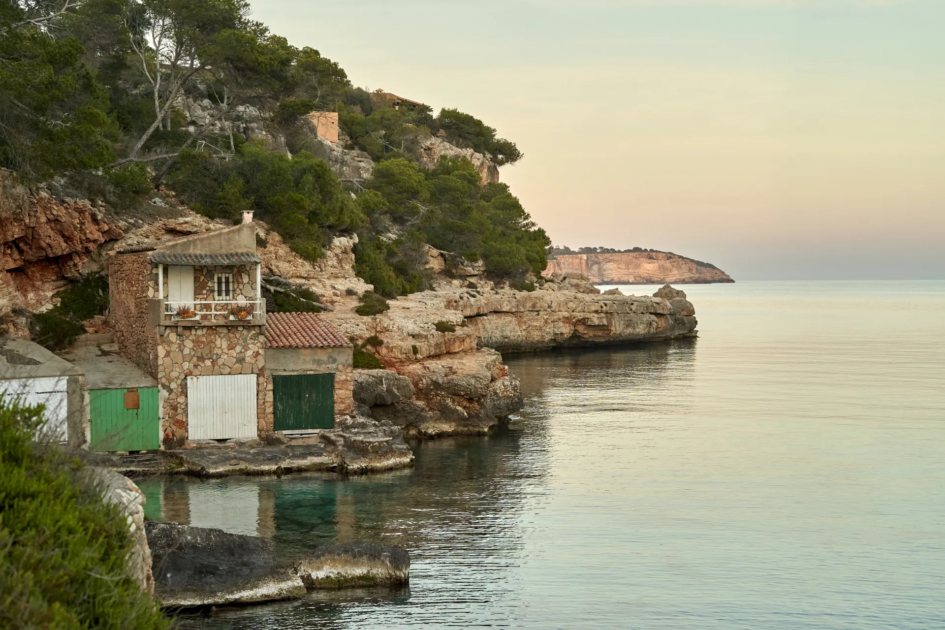 Maisons de pêcheurs traditionnelles en pierre et bois au bord d'une crique rocheuse, mer calme au lever du soleil, végétation méditerranéenne.