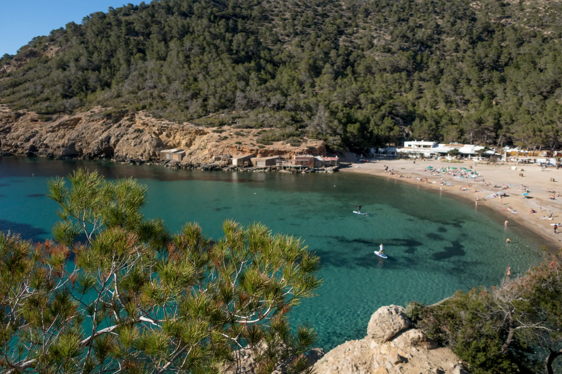 Vue aérienne d'une crique méditerranéenne avec une plage de sable, des eaux turquoise et des collines boisées en arrière-plan sous un ciel bleu.