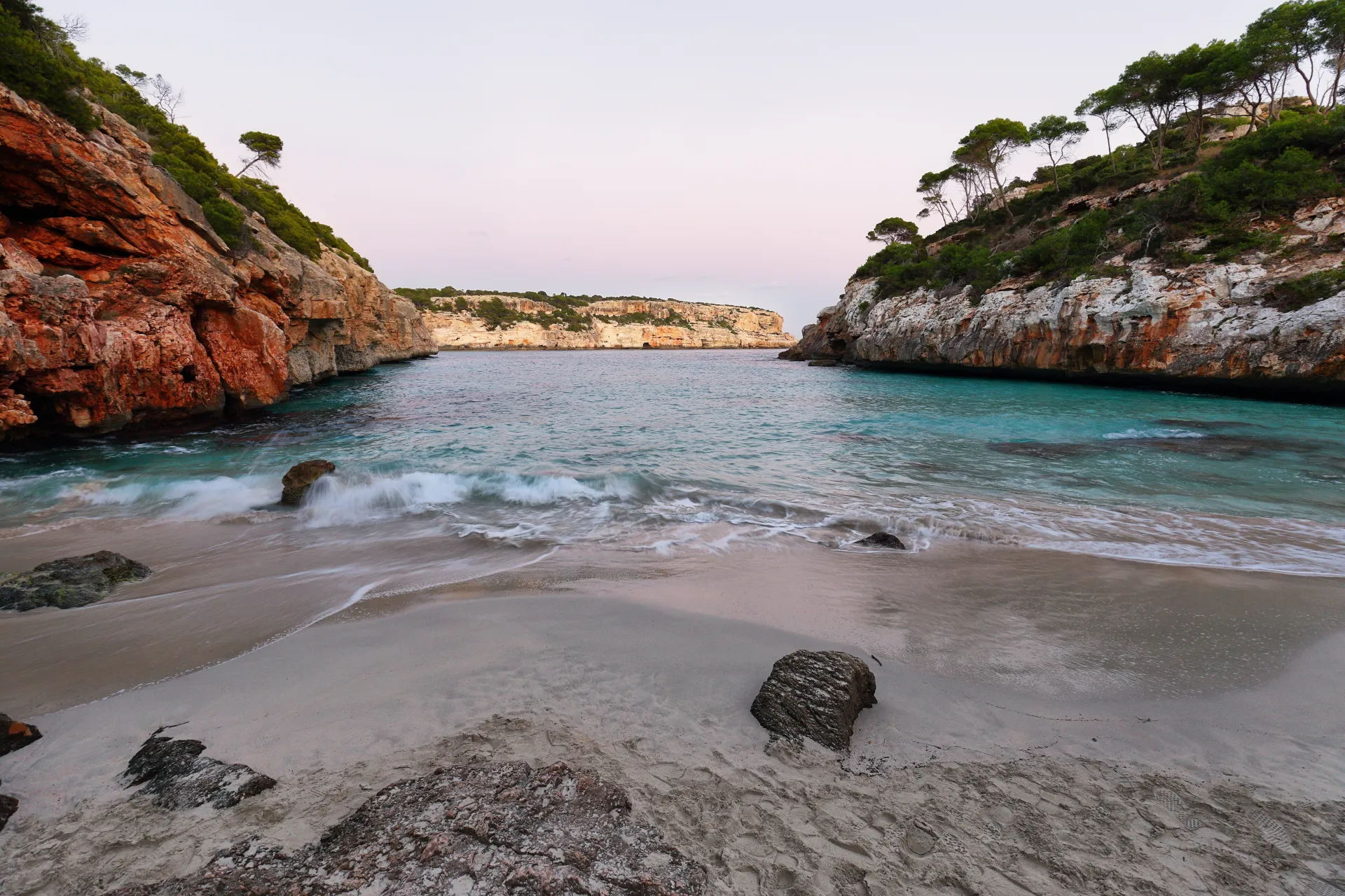 Plage de sable fin avec des vagues turquoise, entourée de falaises rocheuses et de pins méditerranéens sous un ciel clair