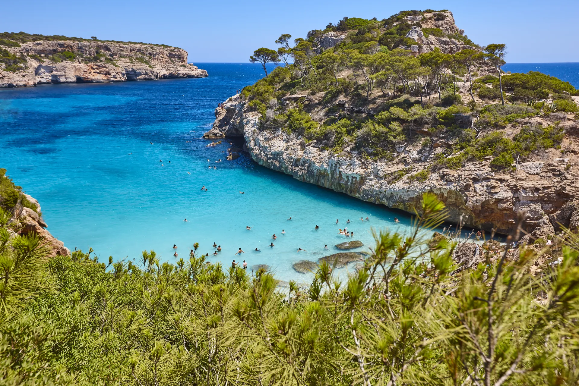 Vue aérienne d'une crique méditerranéenne aux eaux turquoise et cristallines, entourée de falaises rocheuses et de végétation luxuriante, avec des baigneurs
