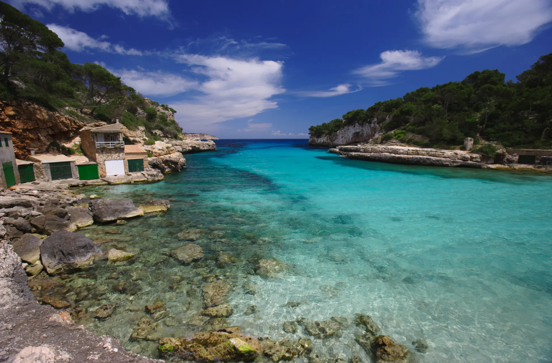 Crique rocheuse aux eaux turquoise et cristallines, avec des cabanes de pêcheurs traditionnelles et une végétation méditerranéenne luxuriante sous un ciel bleu intense.