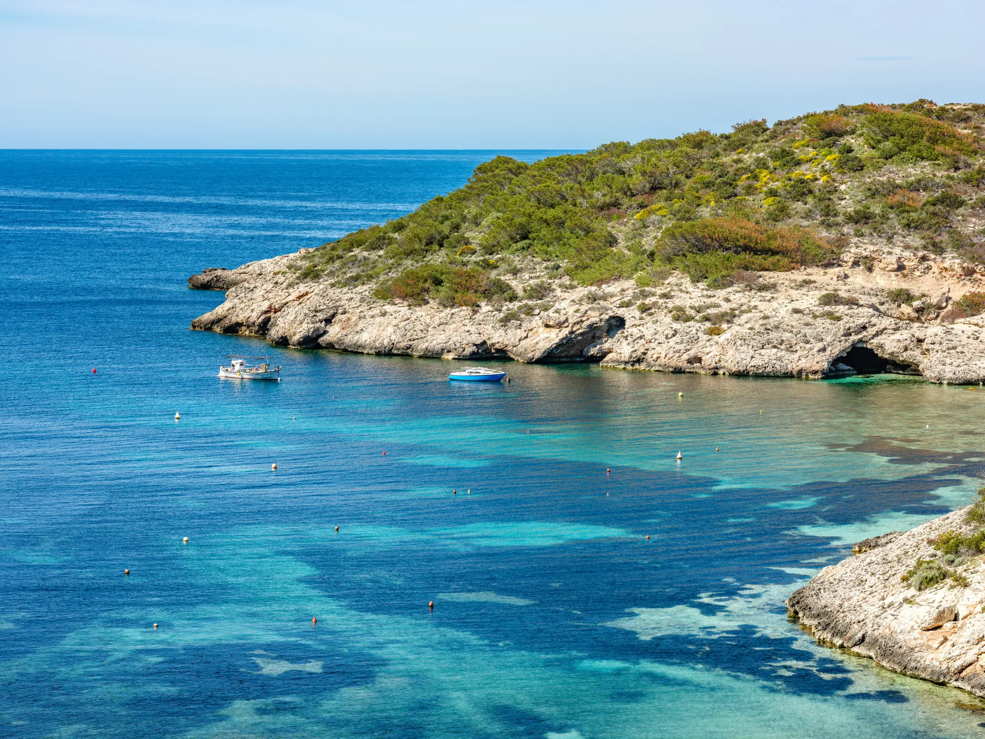 Vue aérienne d'une crique rocheuse aux eaux turquoise et bleues profondes, avec des bateaux ancrés et une végétation méditerranéenne luxuriante sur les falaises.