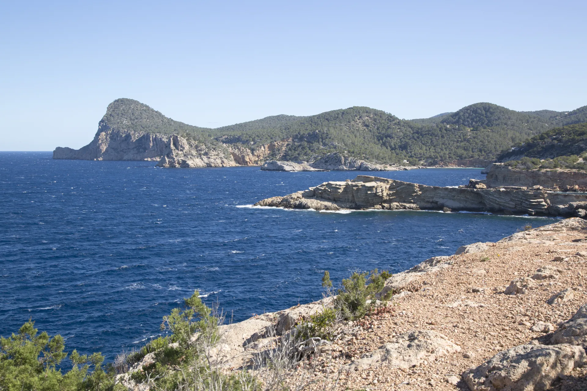 Vue panoramique sur une côte rocheuse méditerranéenne avec des falaises escarpées, une mer bleue et des collines verdoyantes sous un ciel clair