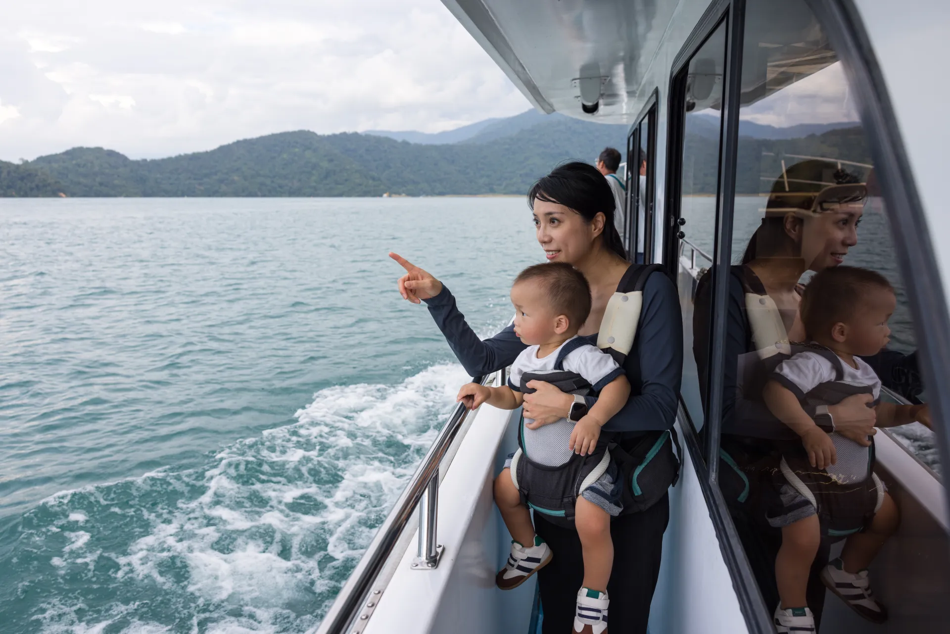 Une mère et son bébé en porte-bébé sur un bateau, la mère pointant du doigt l'horizon marin avec des montagnes verdoyantes en arrière-plan.