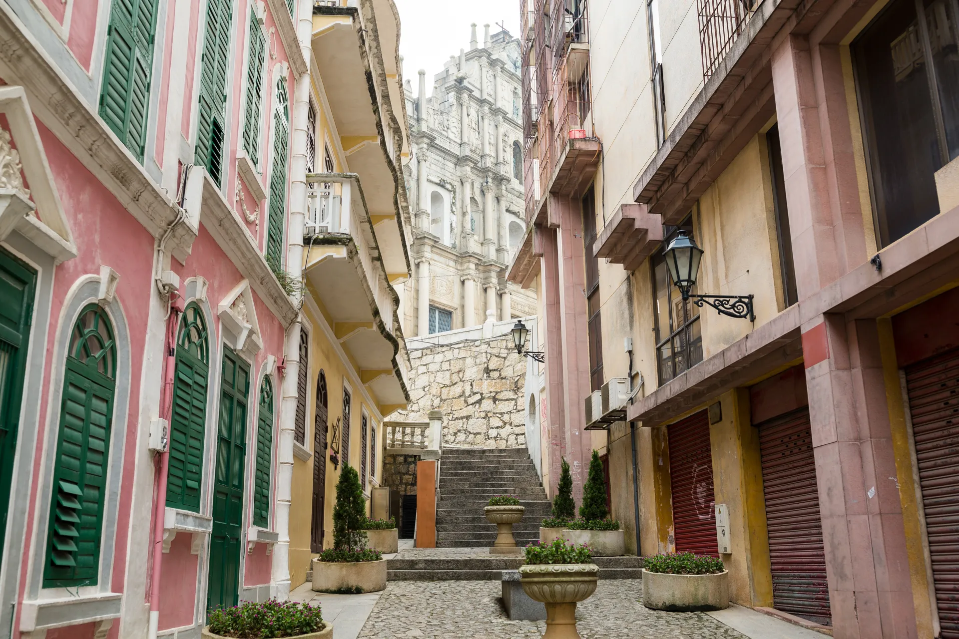 Rue étroite et pavée bordée de bâtiments colorés avec des volets verts, menant à un escalier et un grand bâtiment historique en arrière-plan