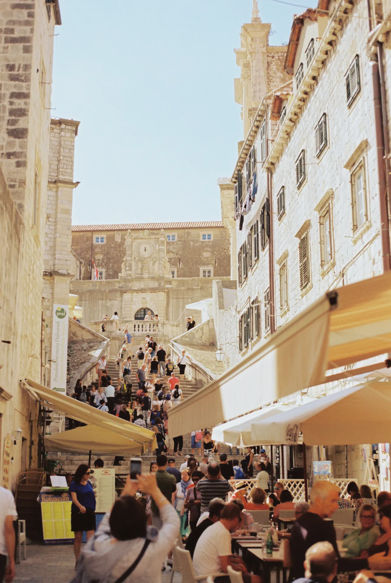 Rue animée d'une ville méditerranéenne avec des escaliers, des bâtiments en pierre et des terrasses de cafés bondées sous le soleil.