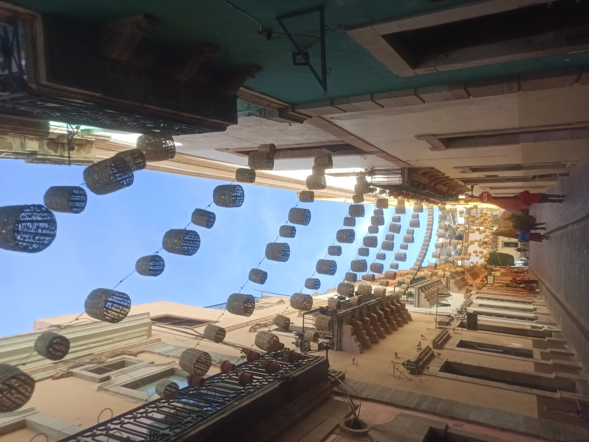 Vue en contre-plongée d'une rue étroite décorée de guirlandes de paniers suspendus, avec des façades de bâtiments anciens et un ciel bleu clair