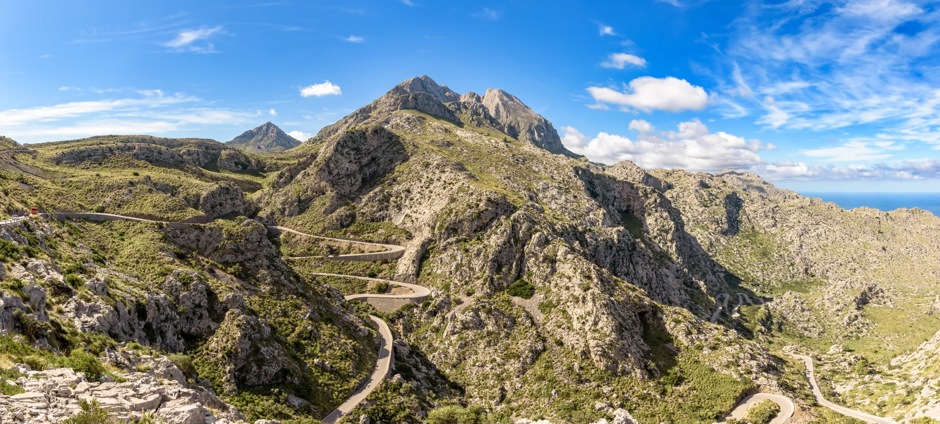 Vue panoramique d'une route sinueuse de montagne serpentant à travers des paysages rocheux et verdoyants sous un ciel bleu parsemé de nuages blancs