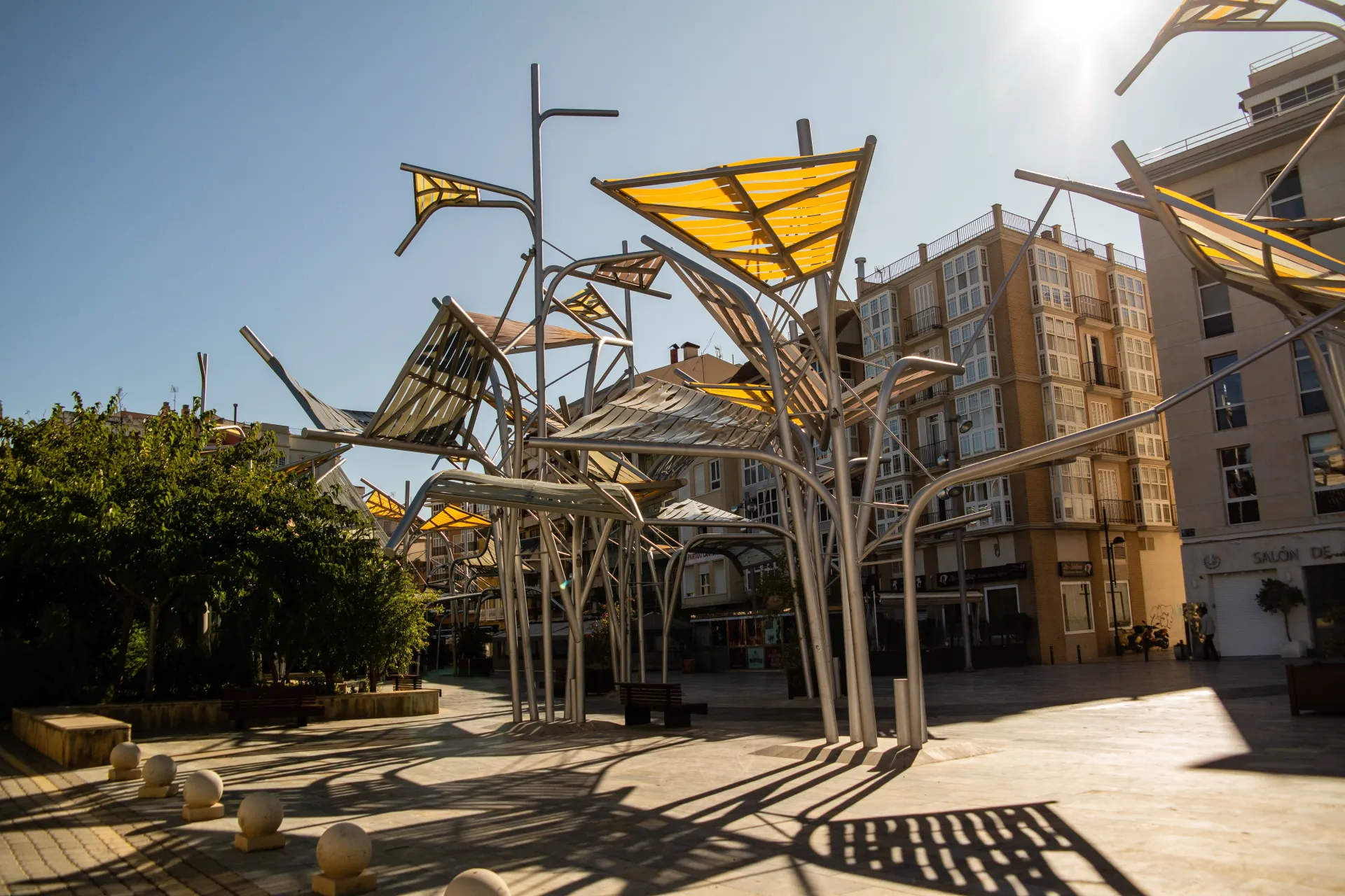 Une place publique moderne avec des structures métalliques jaunes et grises offrant de l'ombre, des arbres verts et des bâtiments en arrière-plan sous un ciel bleu clair.