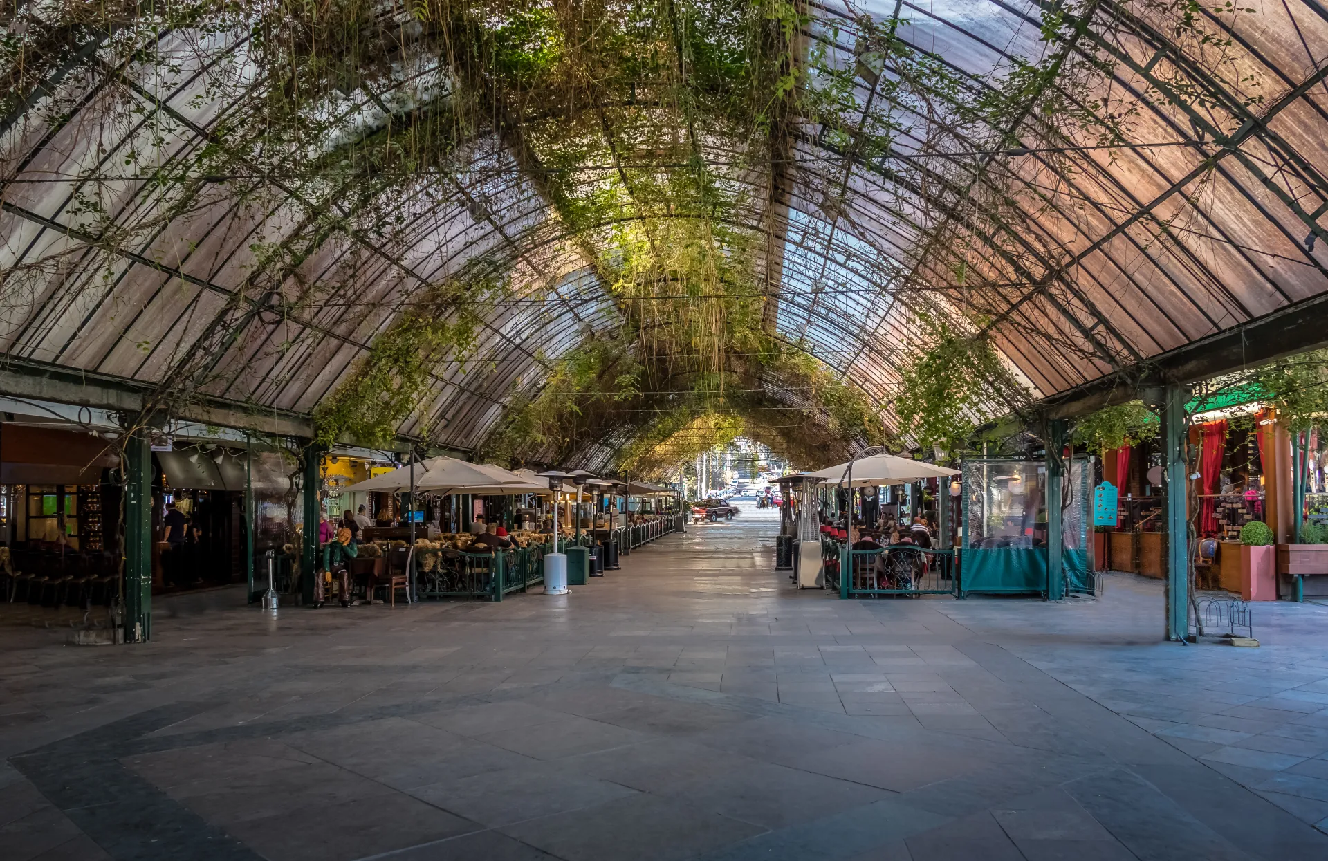 Allée couverte d'une structure métallique et vitrée, ornée de plantes grimpantes, avec des terrasses de restaurants et des parasols