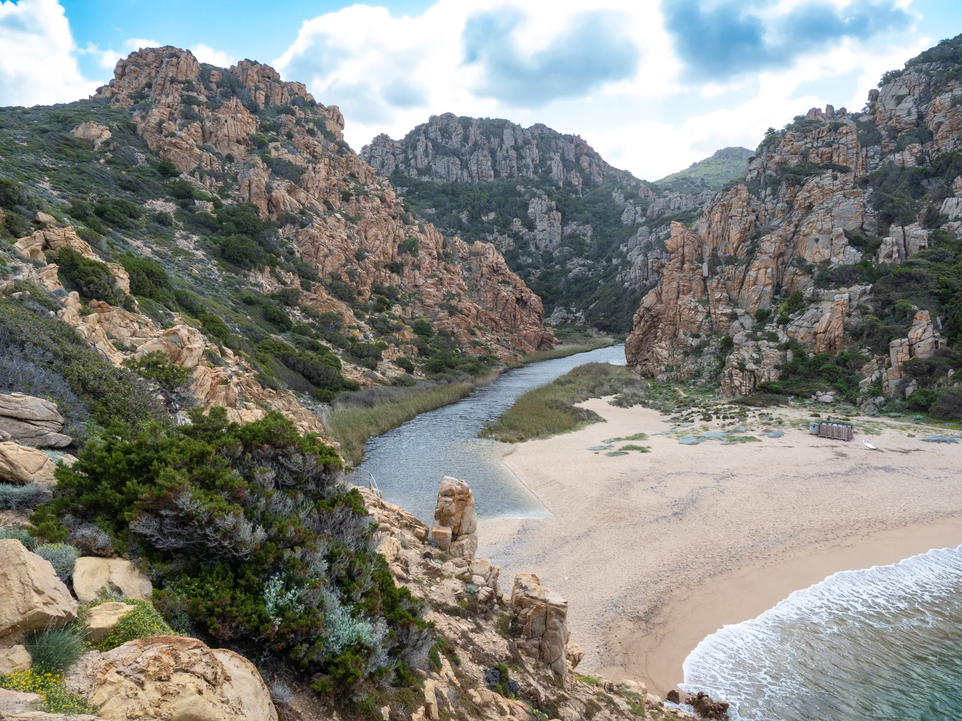 Vue aérienne d'une plage de sable fin où une rivière se jette dans la mer, entourée de falaises rocheuses et de végétation méditerranéenne sous un ciel nuageux.