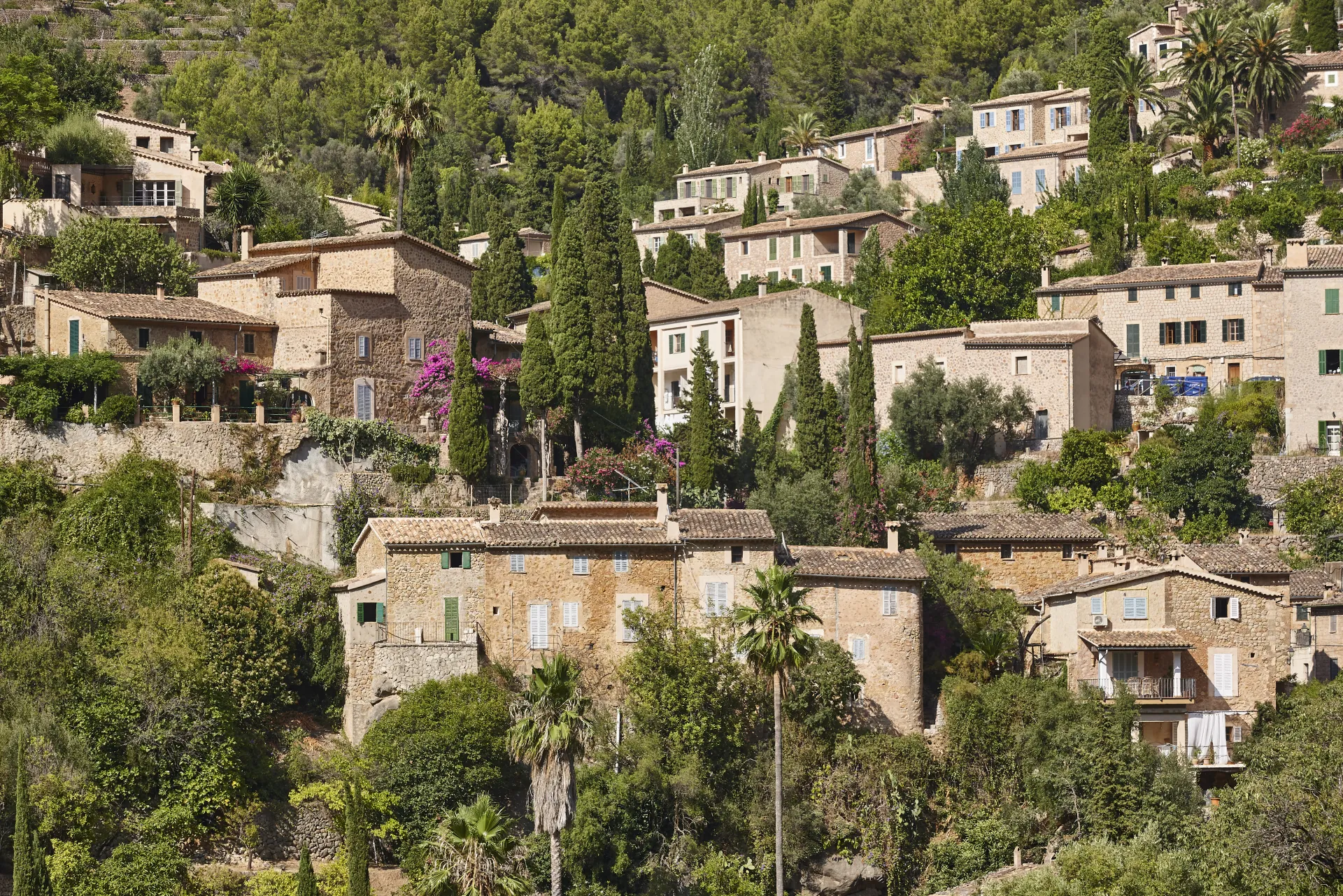 Village méditerranéen en pierre sur une colline verdoyante, maisons traditionnelles avec toits de tuiles et végétation luxuriante, ambiance paisible.