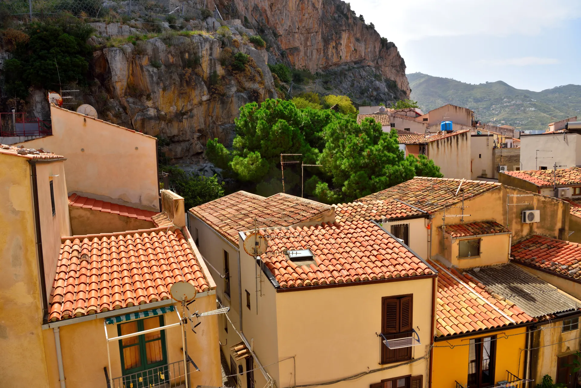 Vue plongeante sur des toits de tuiles oranges d'un village méditerranéen, avec des maisons colorées et une montagne rocheuse en arrière-plan sous un ciel bleu.