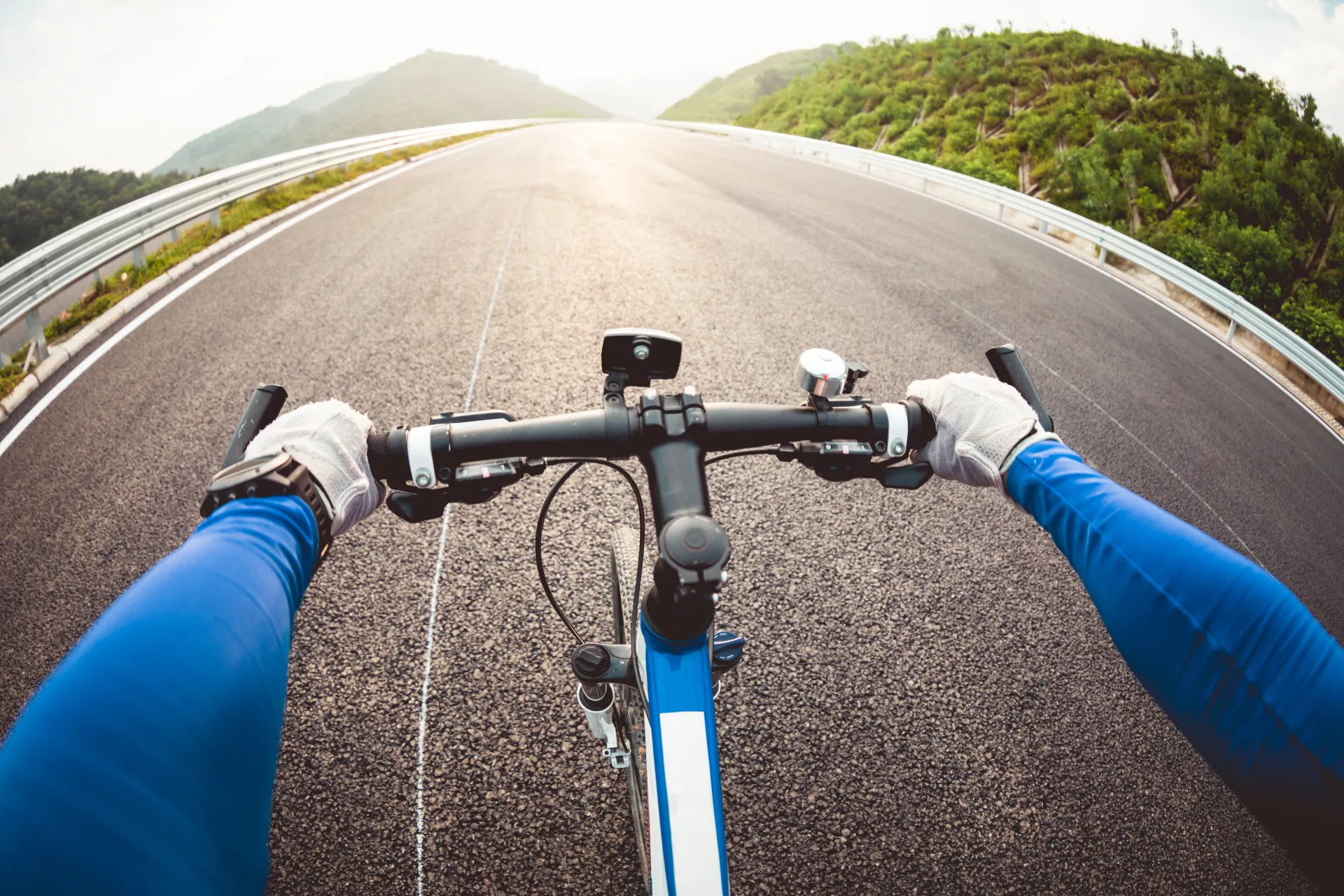 Vue à la première personne d'un cycliste sur une route goudronnée sinueuse avec des montagnes verdoyantes en arrière-plan sous un ciel lumineux.