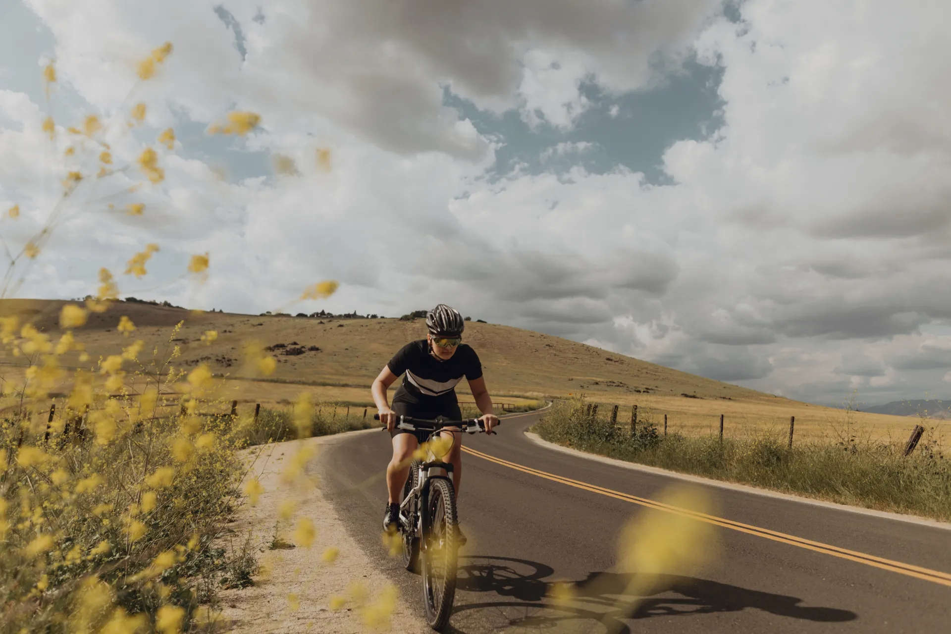 Cycliste sur un vélo de route, portant un casque et des lunettes de soleil, sur une route sinueuse bordée de champs jaunes et de collines sous un ciel nuageux.