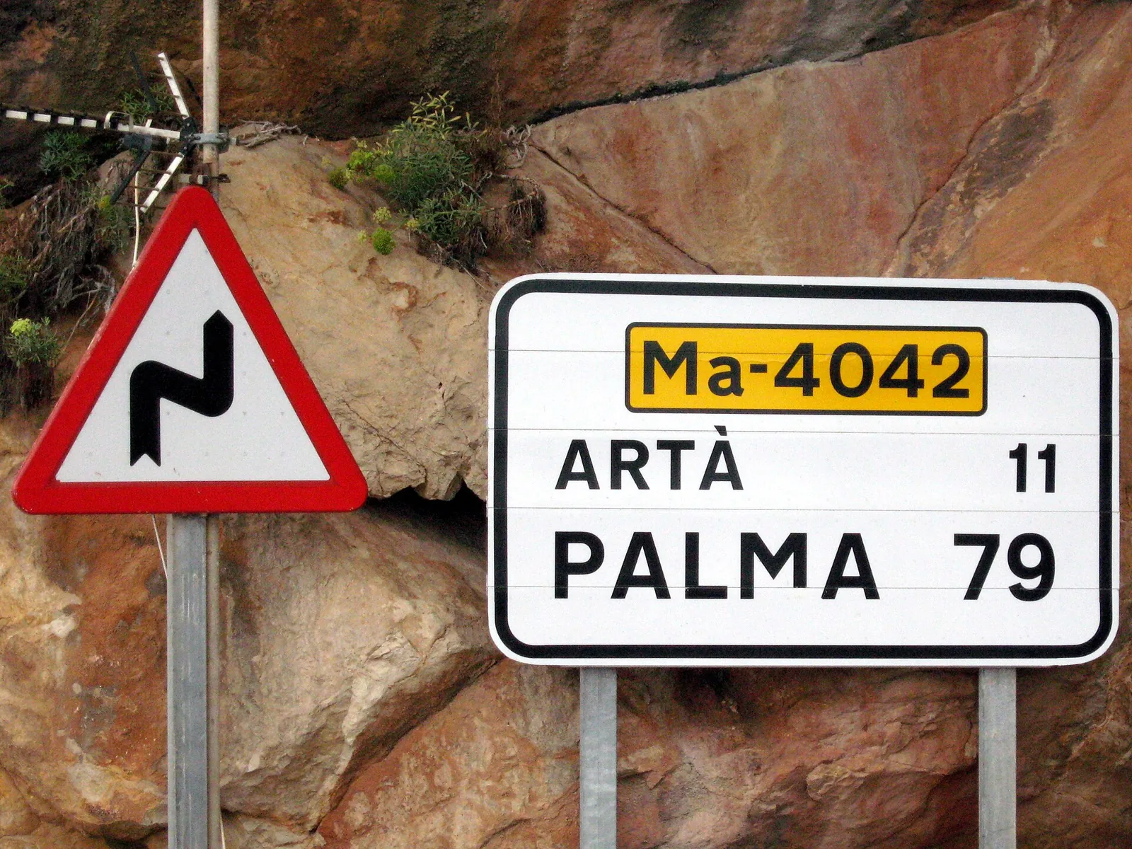 Panneau de la route Ma-4042 (Artà 11, Palma 79) à côté d’un panneau de virages, devant une paroi de roche rouge